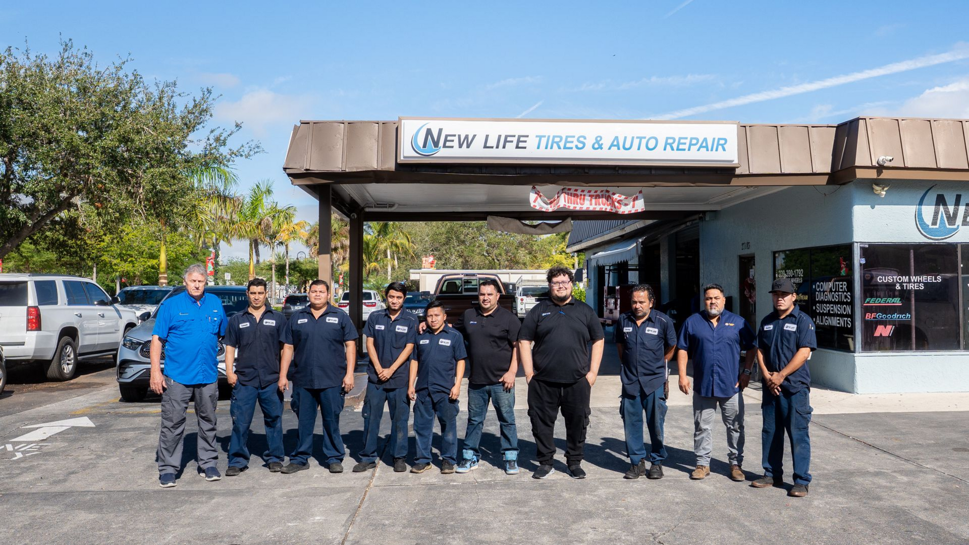 Group of auto repair shop employees stand in front of the shop. Blue shirts, smiling. Sign: