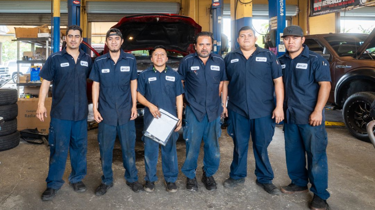 Six auto mechanics in blue uniforms standing in a garage, in front of a car with its hood open.