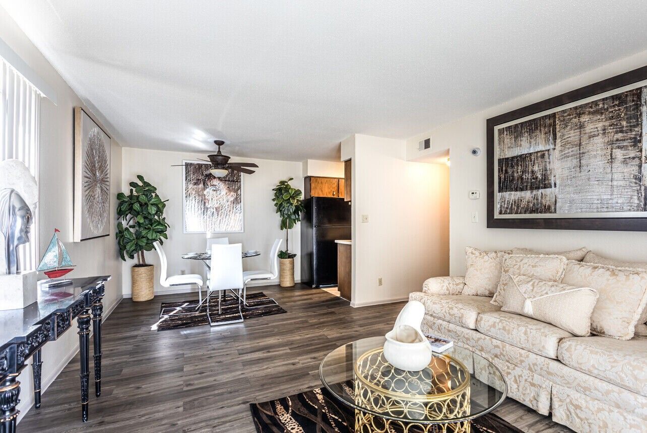Living room with sofa, table, and dining area. Neutral colors with modern decor and artificial plants.