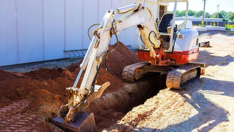 A white and red excavator digging a trench in the reddish-brown earth at a construction site.
