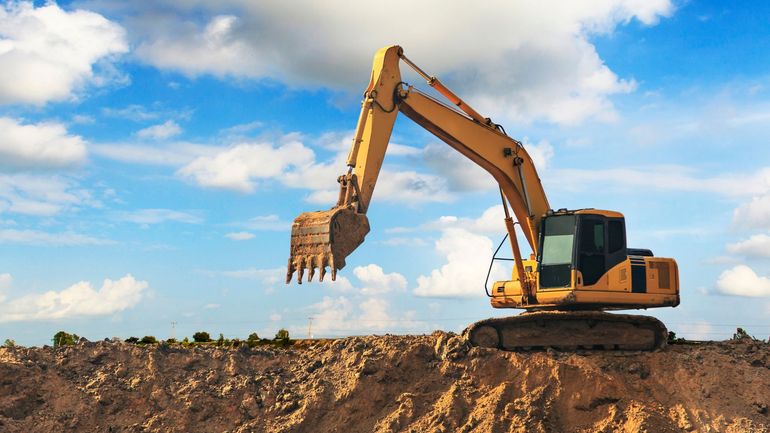 A yellow excavator positioned on top of a rocky mound against a bright blue, cloudy sky.