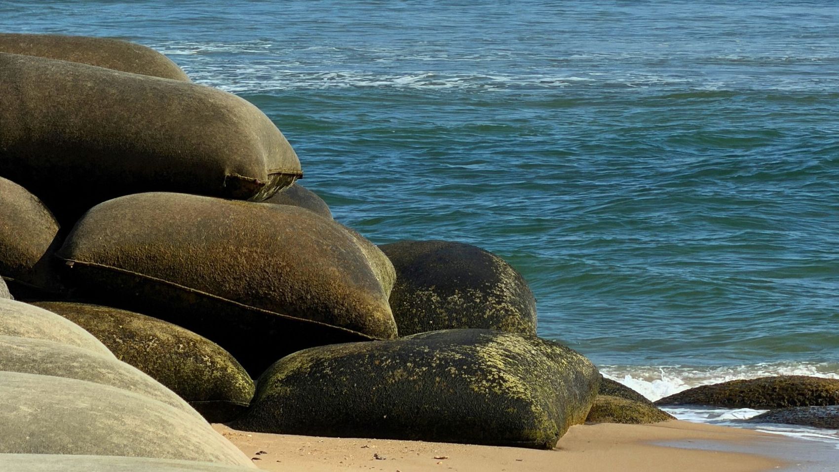 Large, mossy, dark gray sandbags stacked on a sandy beach next to the ocean.