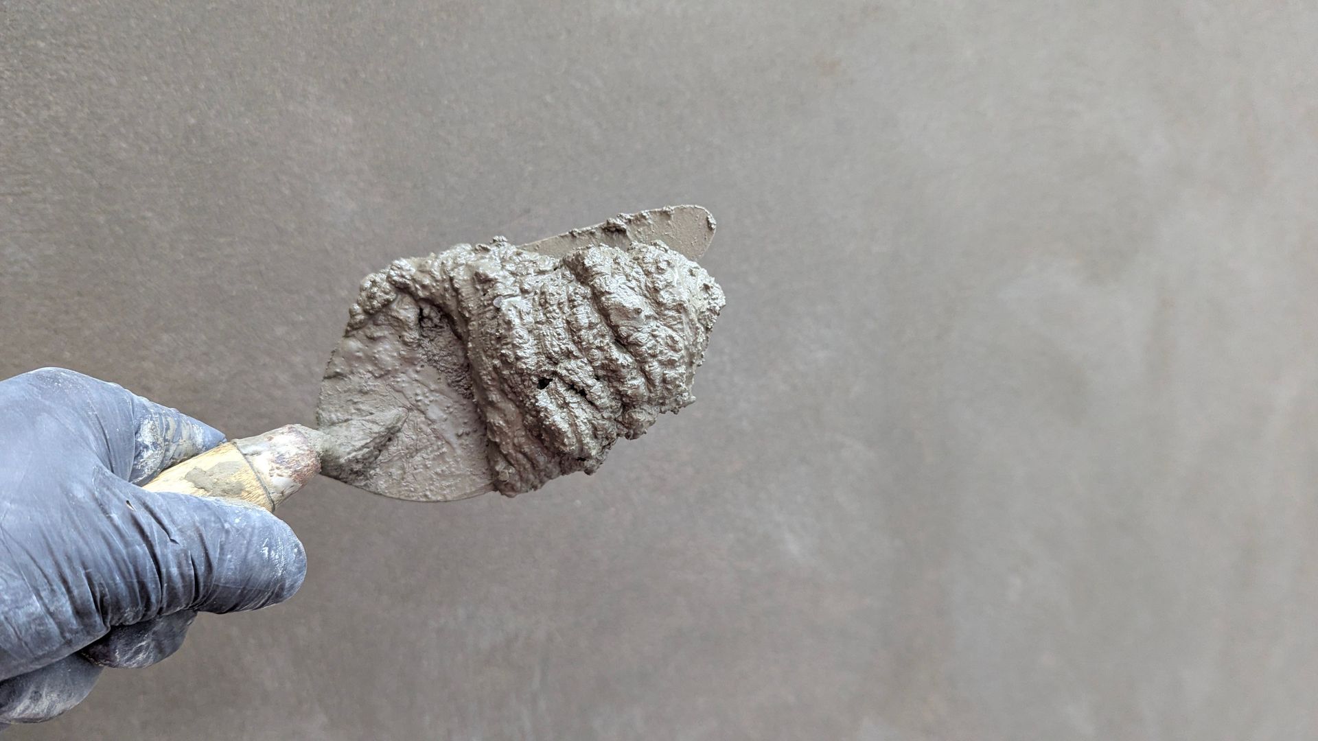 Gloved hand holding a small trowel with gray plaster against a smooth gray wall