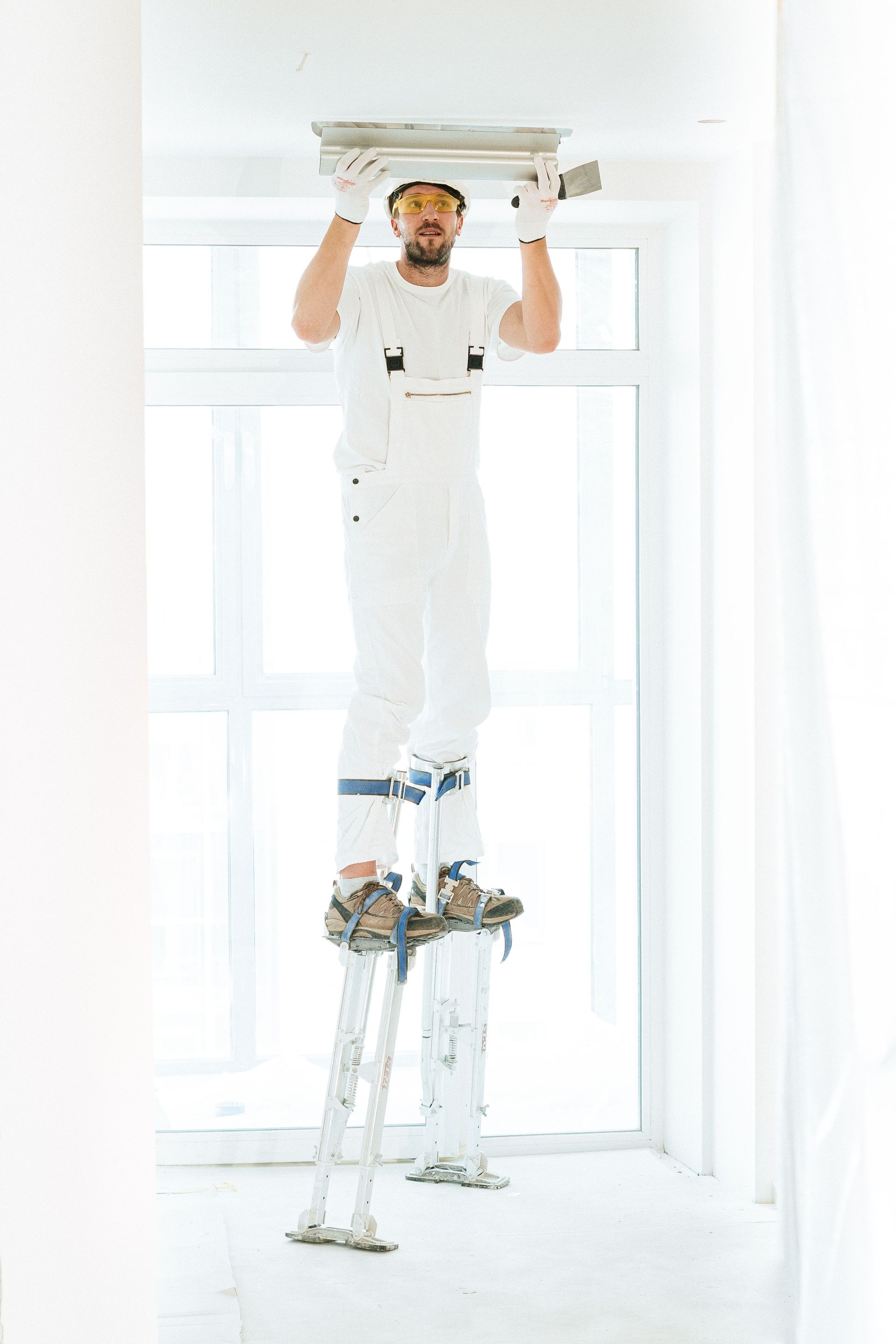 Painter standing on a ladder, installing or painting a ceiling fixture in a bright white room.