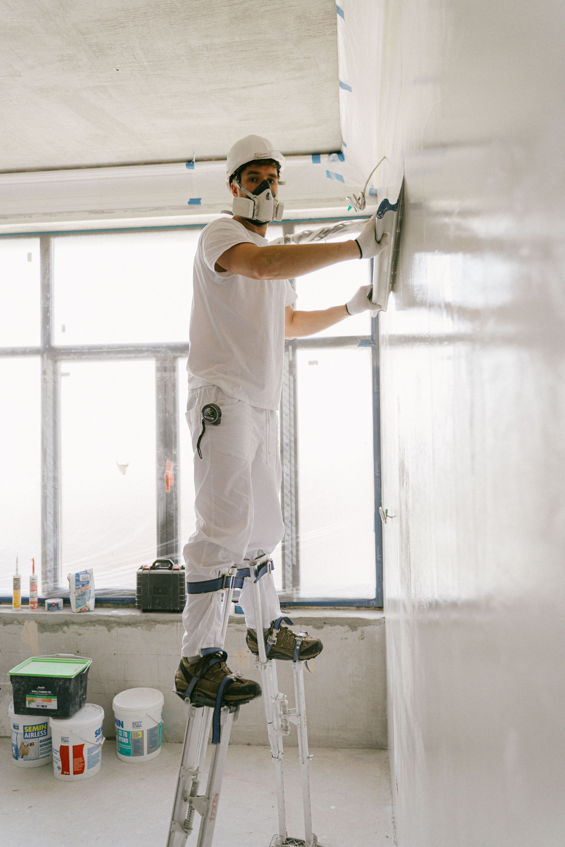 Painter on stilts painting a white wall in a bright, unfinished room