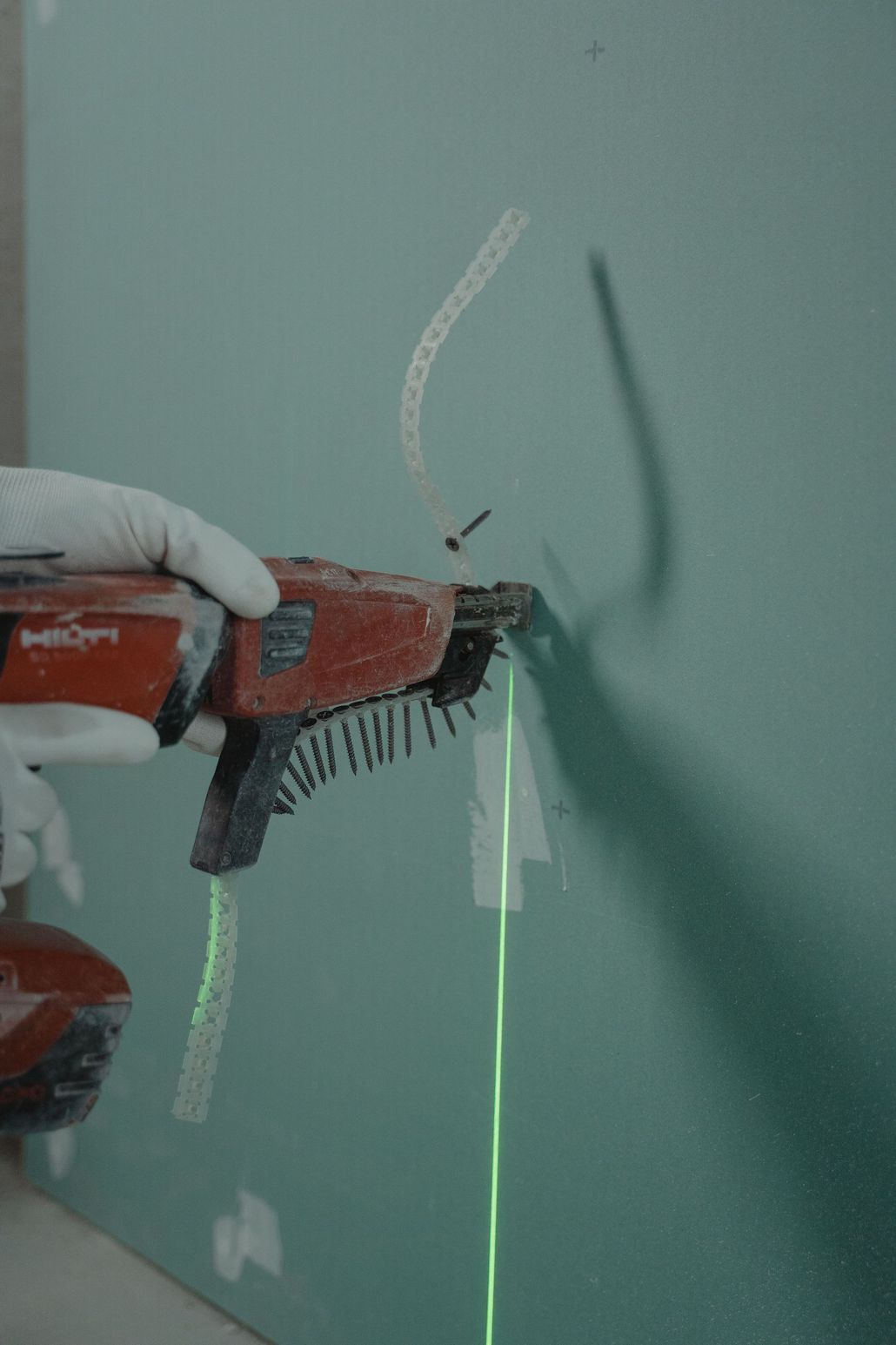 A gloved hand drills a hole in a green wall, with dust marking the drilling spot.