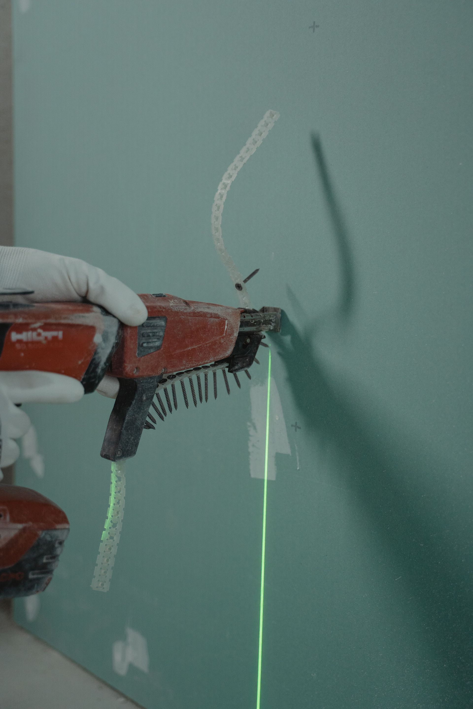 A person uses a red power drill against a green wall, creating a shadow and a small mark.
