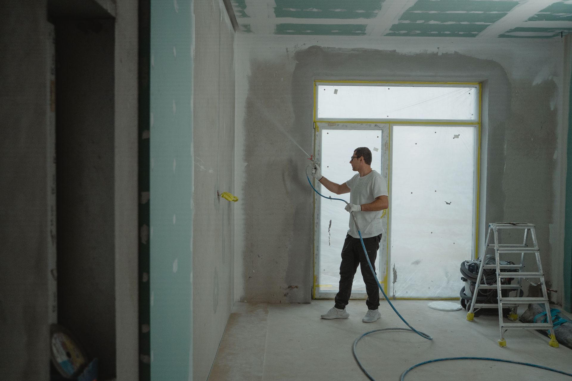 Person painting drywall in a room under renovation, with ladder and tools nearby.