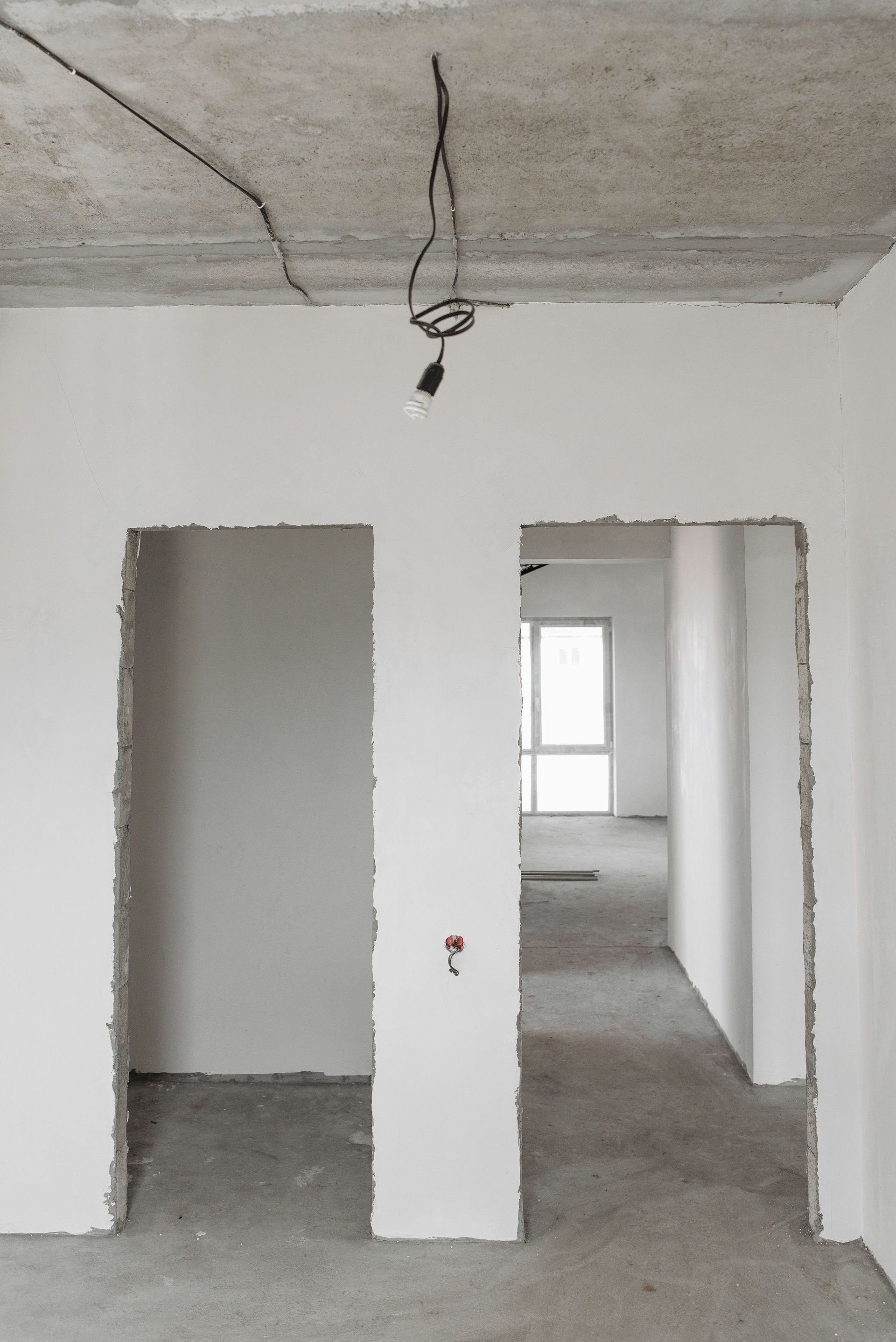 Unfinished apartment interior with white walls, exposed ceiling wiring, and open doorways leading to a bright room.