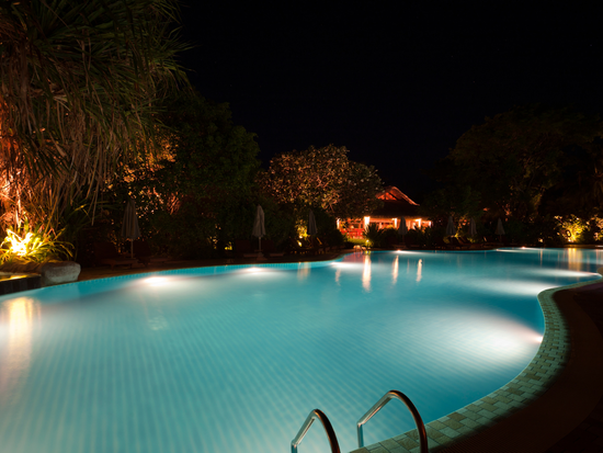 Nighttime view of a turquoise pool lit by underwater lights, with surrounding trees and a building in the distance.