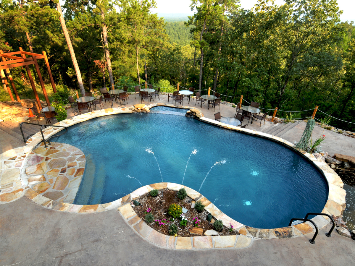 Pool with a stone border and fountains, surrounded by trees and tables for dining.