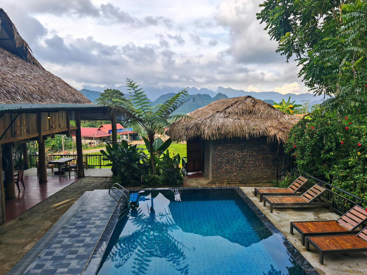 Swimming pool and thatched-roof buildings with mountain backdrop. Lounge chairs beside pool. Cloudy sky.
