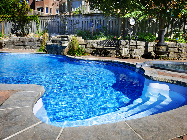 Blue swimming pool with stone steps and waterfall feature in a backyard.