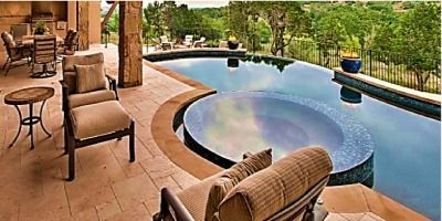 Patio with lounge chairs, round table, dining set, and an infinity pool overlooking a wooded landscape.