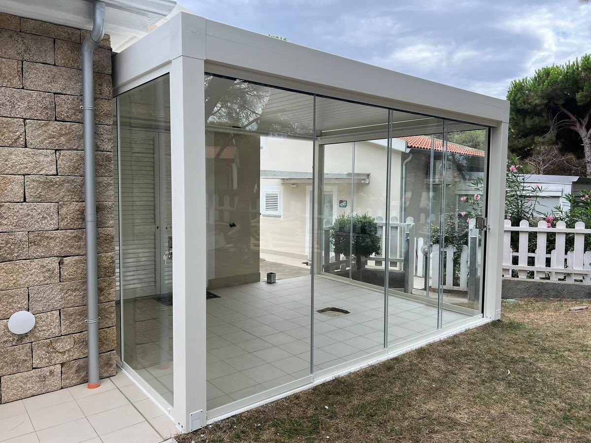 Una moderna struttura in vetro con cornice bianca, annessa a un edificio con muri in pietra, situata su un patio con un prato.