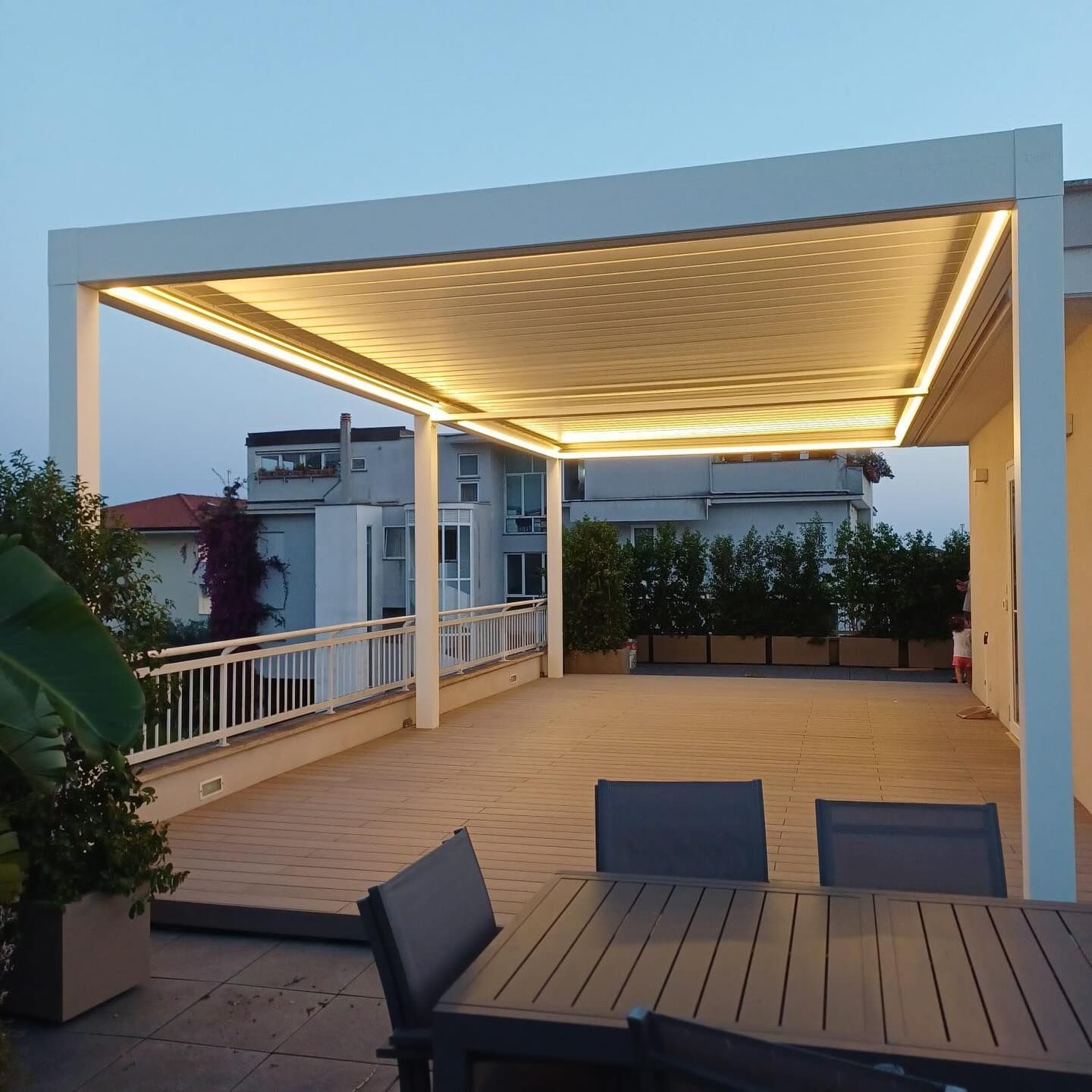 Una pergola bianca con luci LED a luce calda sopra un patio con un set da pranzo in legno su un balcone al crepuscolo.