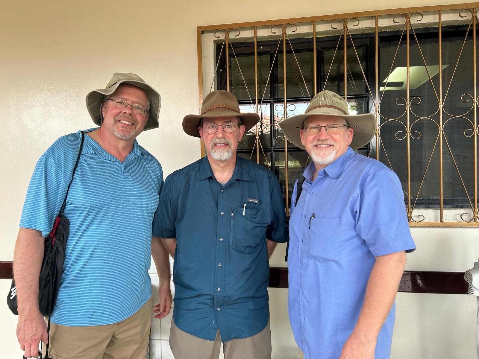 Three men wearing hats are posing for a picture in front of a window