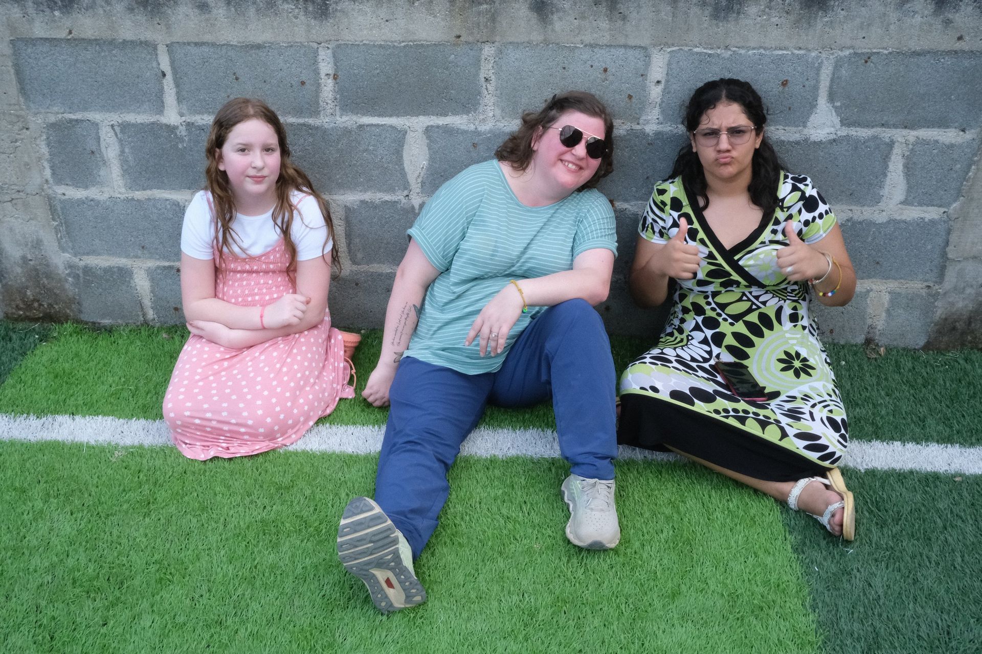 Three women are sitting on the grass in front of a brick wall.
