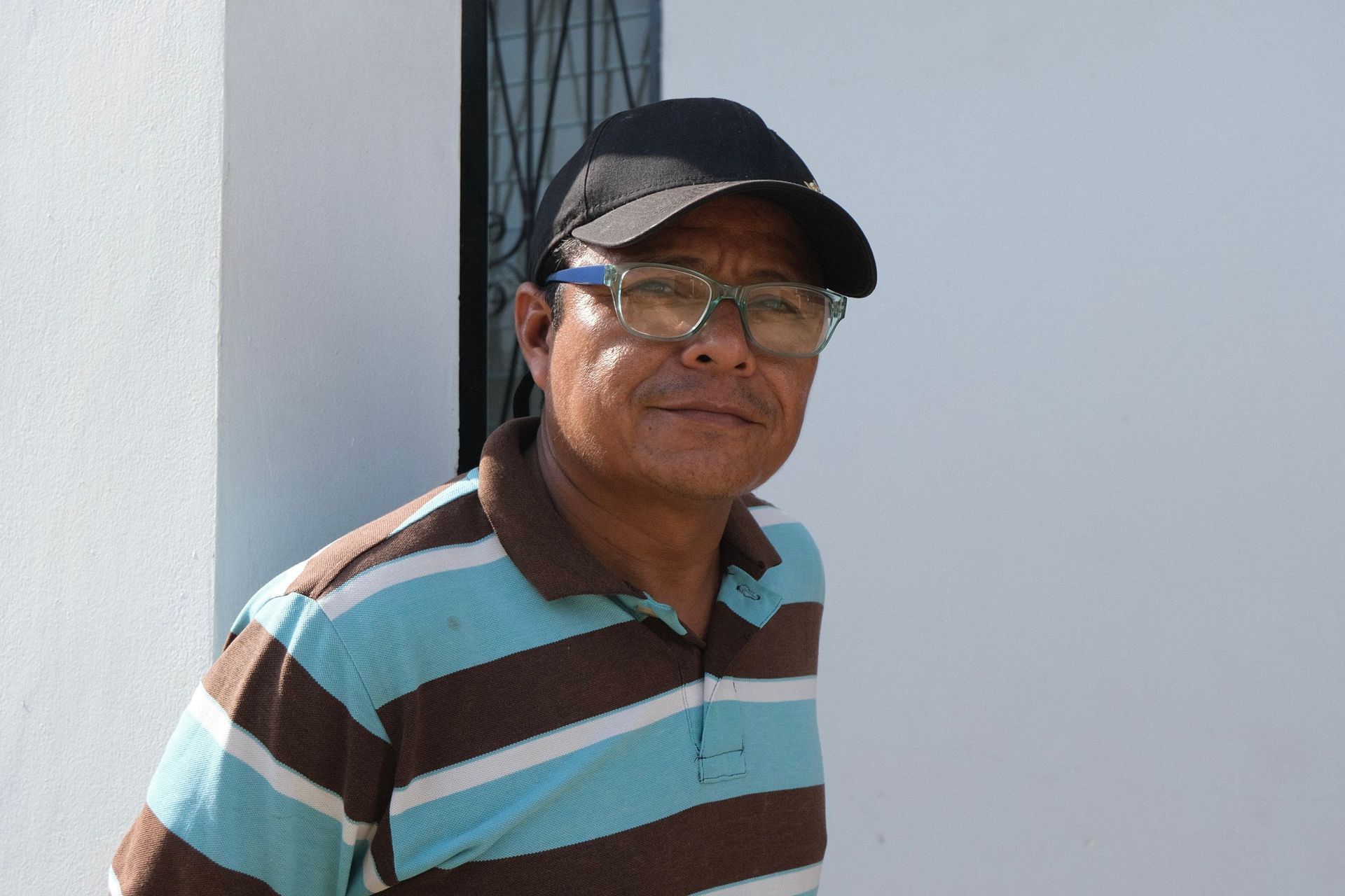 A man wearing glasses and a hat is standing in front of a white wall.