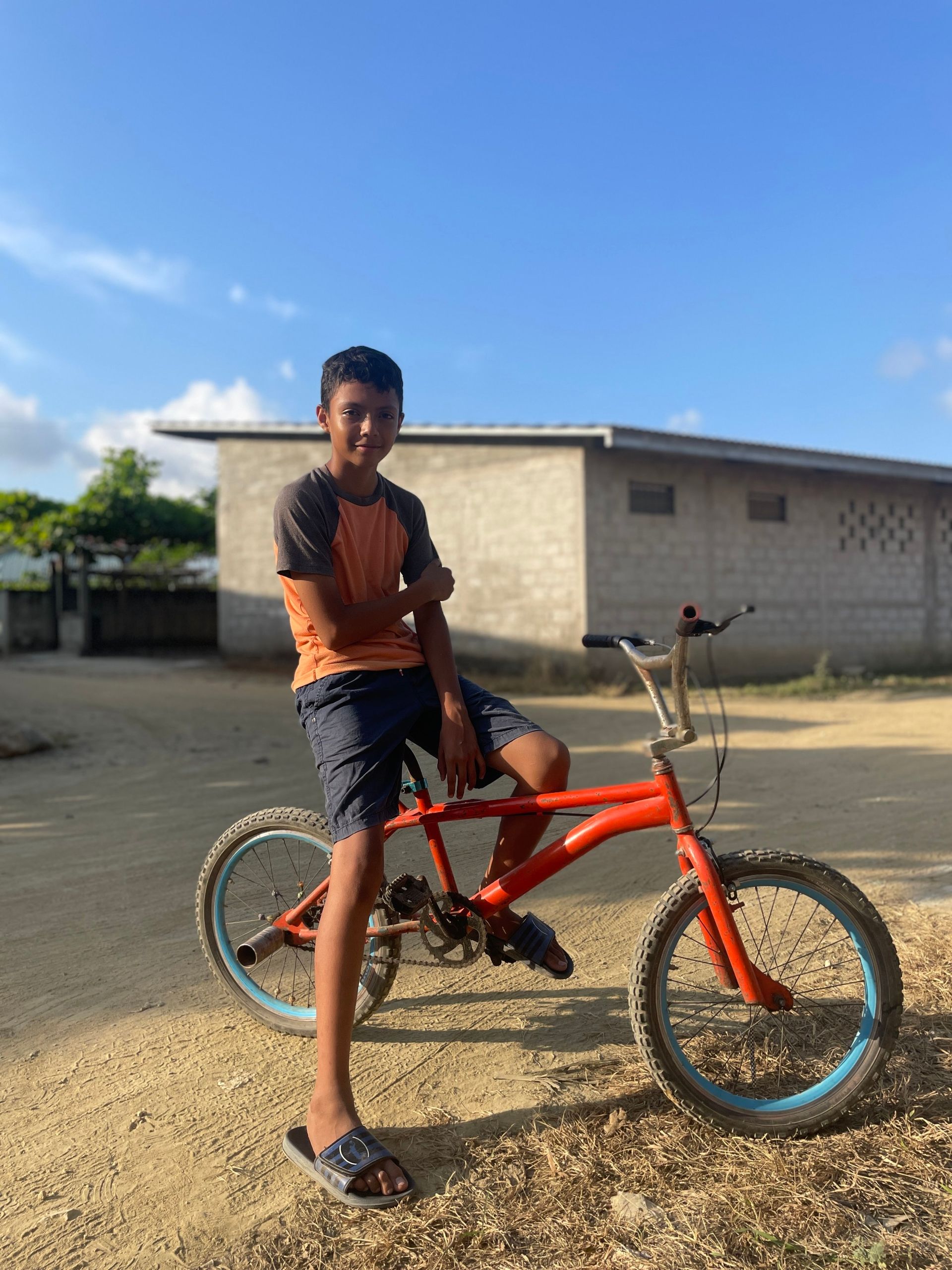 A young boy is sitting on an orange bicycle in a dirt field.