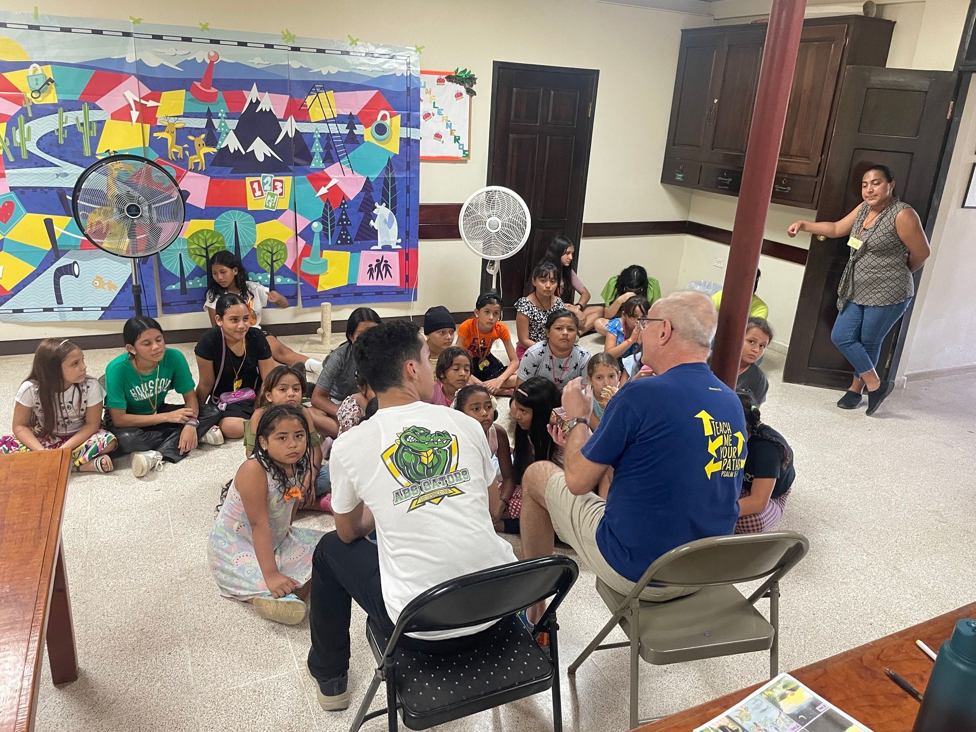 A group of children are sitting on the floor in a room with a man sitting in a chair talking to them.
