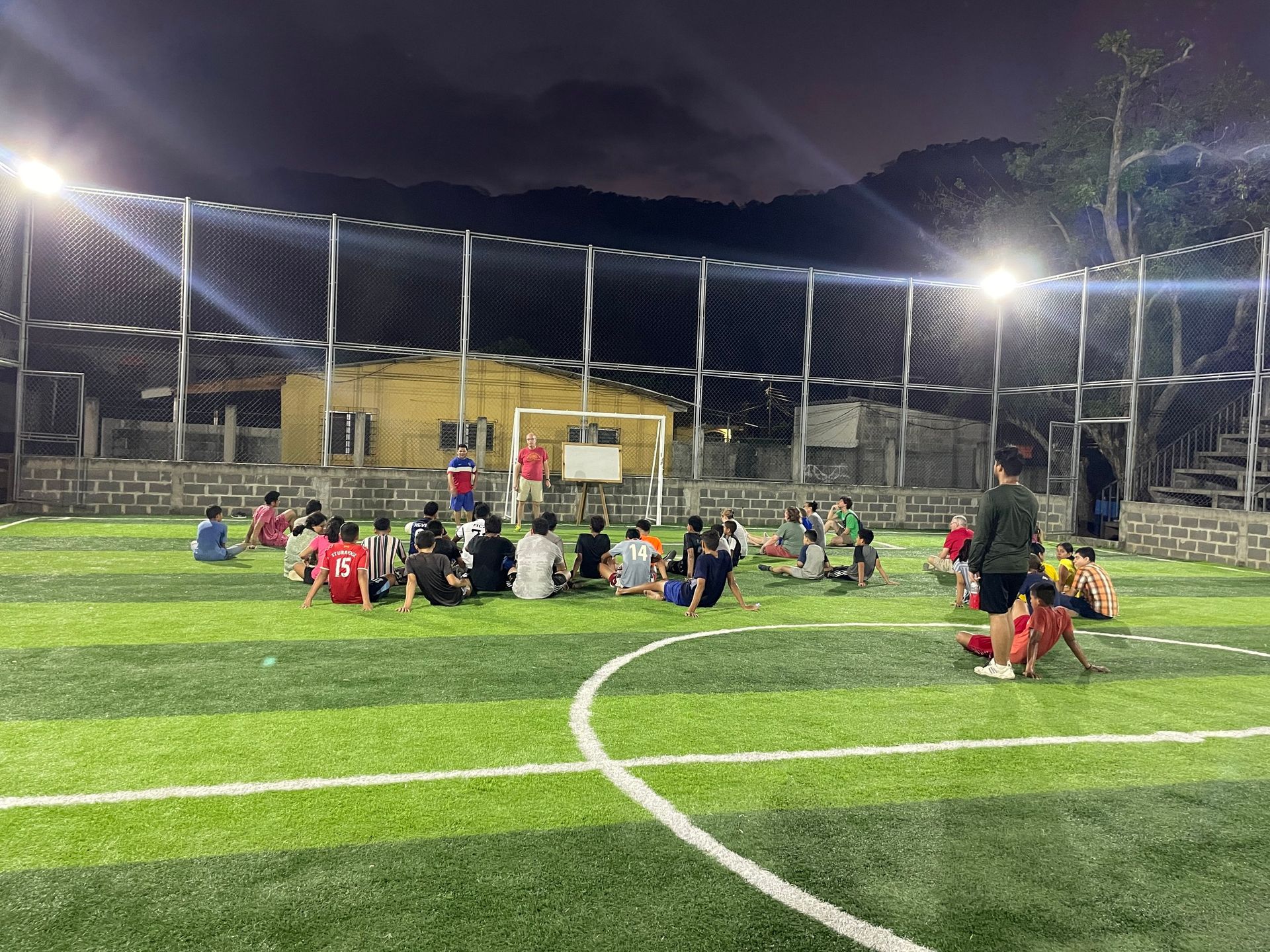 A group of people are sitting on top of a soccer field at night.