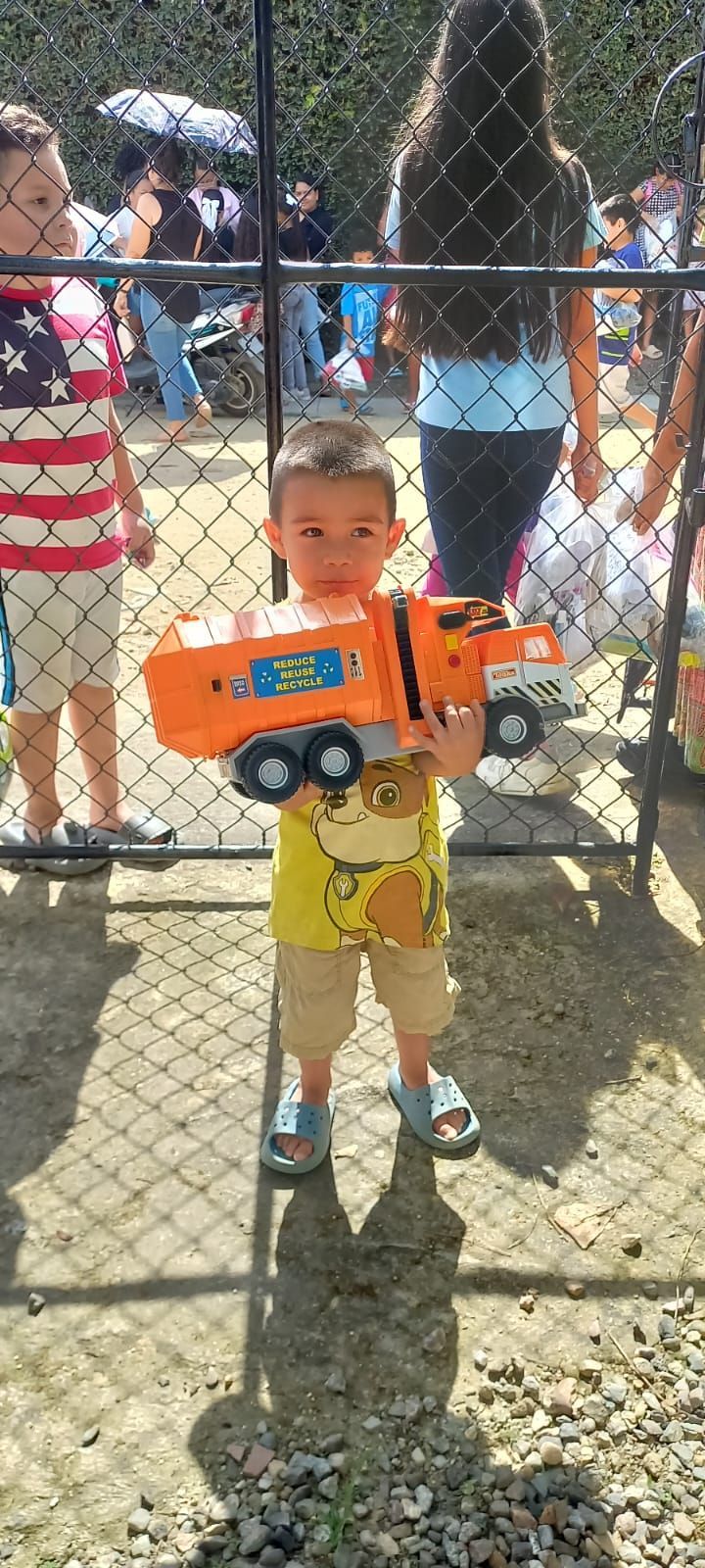 A little boy is holding a toy truck in front of a chain link fence.