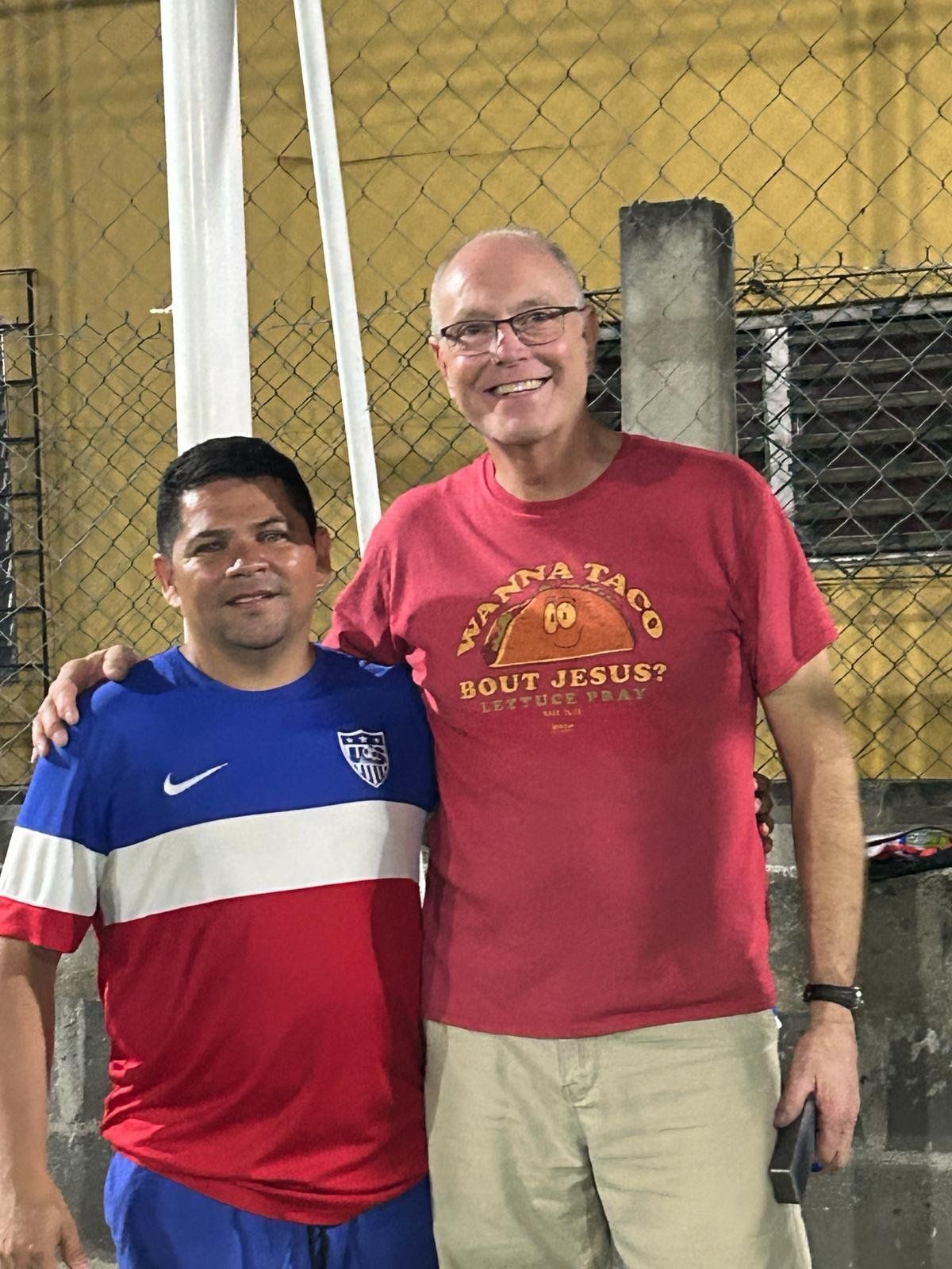 Two men are posing for a picture in front of a chain link fence.