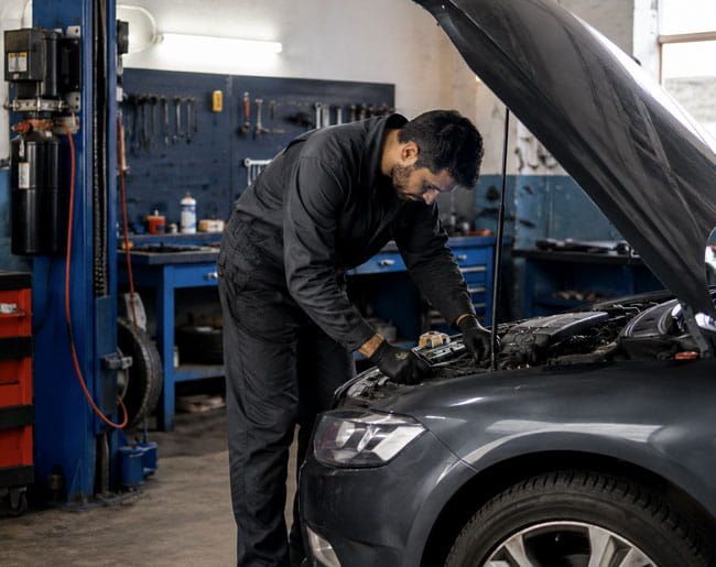 Mecánico trabajando bajo el capó de un SUV gris en un taller de reparación de automóviles.