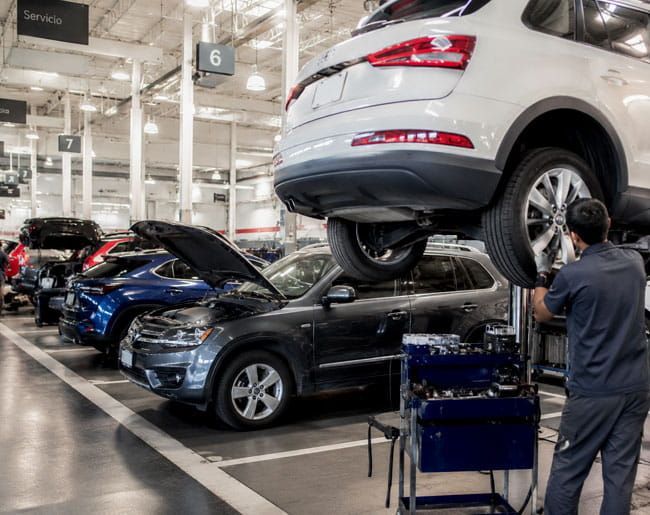 Varios coches apilados en un taller mecánico, con un mecánico inspeccionando un SUV blanco elevado.