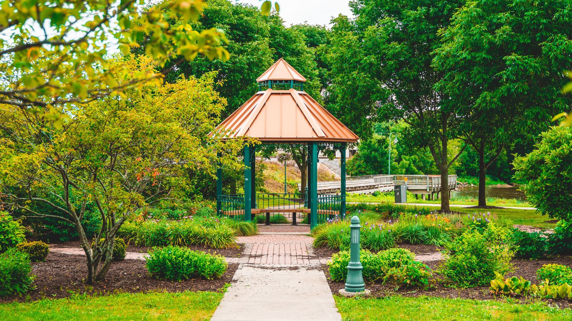 Park gazebo with a red roof surrounded by lush green trees and a paved path leading to it.