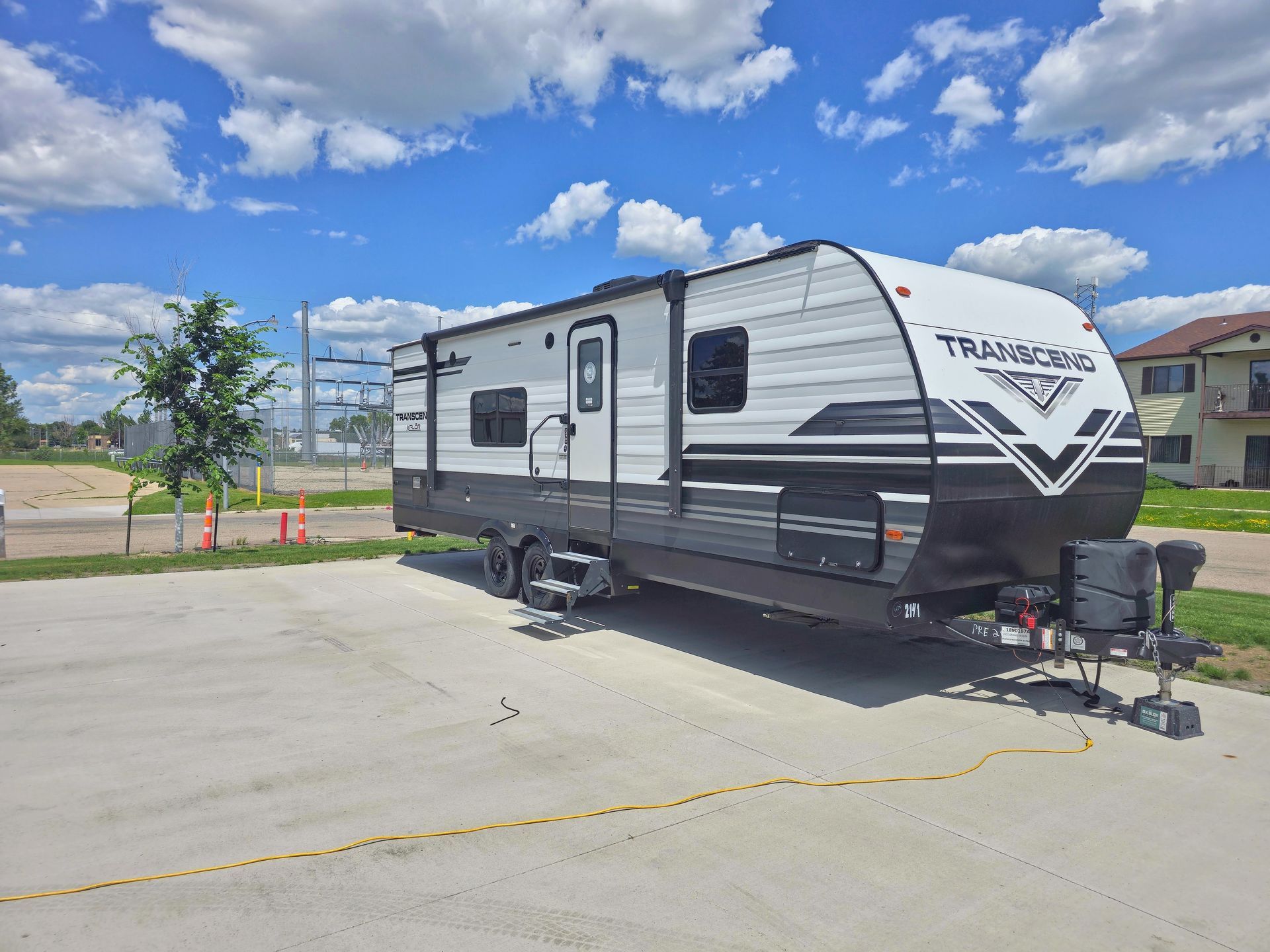 White and gray travel trailer parked on a concrete lot under a blue sky with clouds.