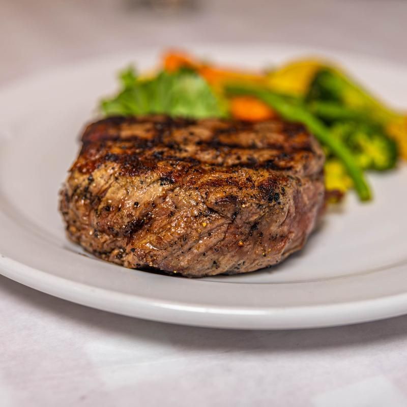 Grilled steak medallion on a white plate with roasted vegetables in the background