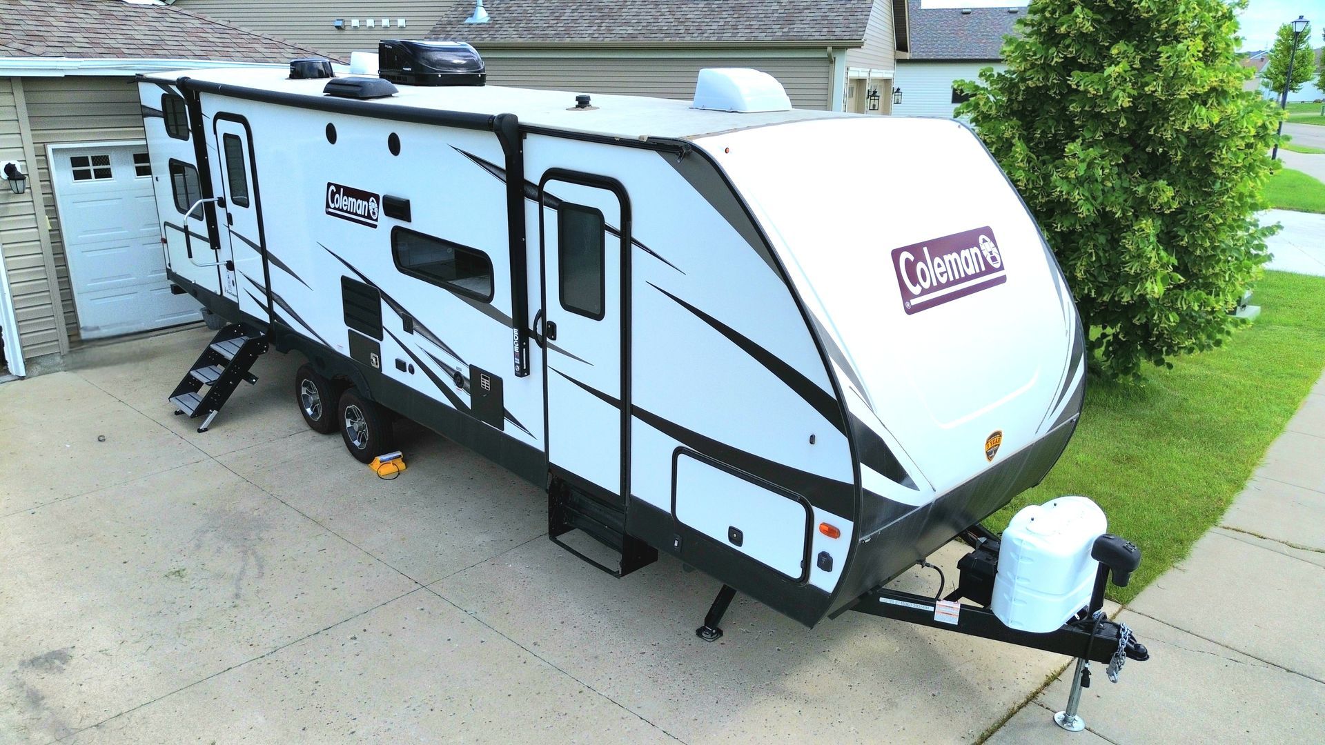 White Coleman travel trailer parked in a driveway beside a house, with steps, awning, and front storage tank.