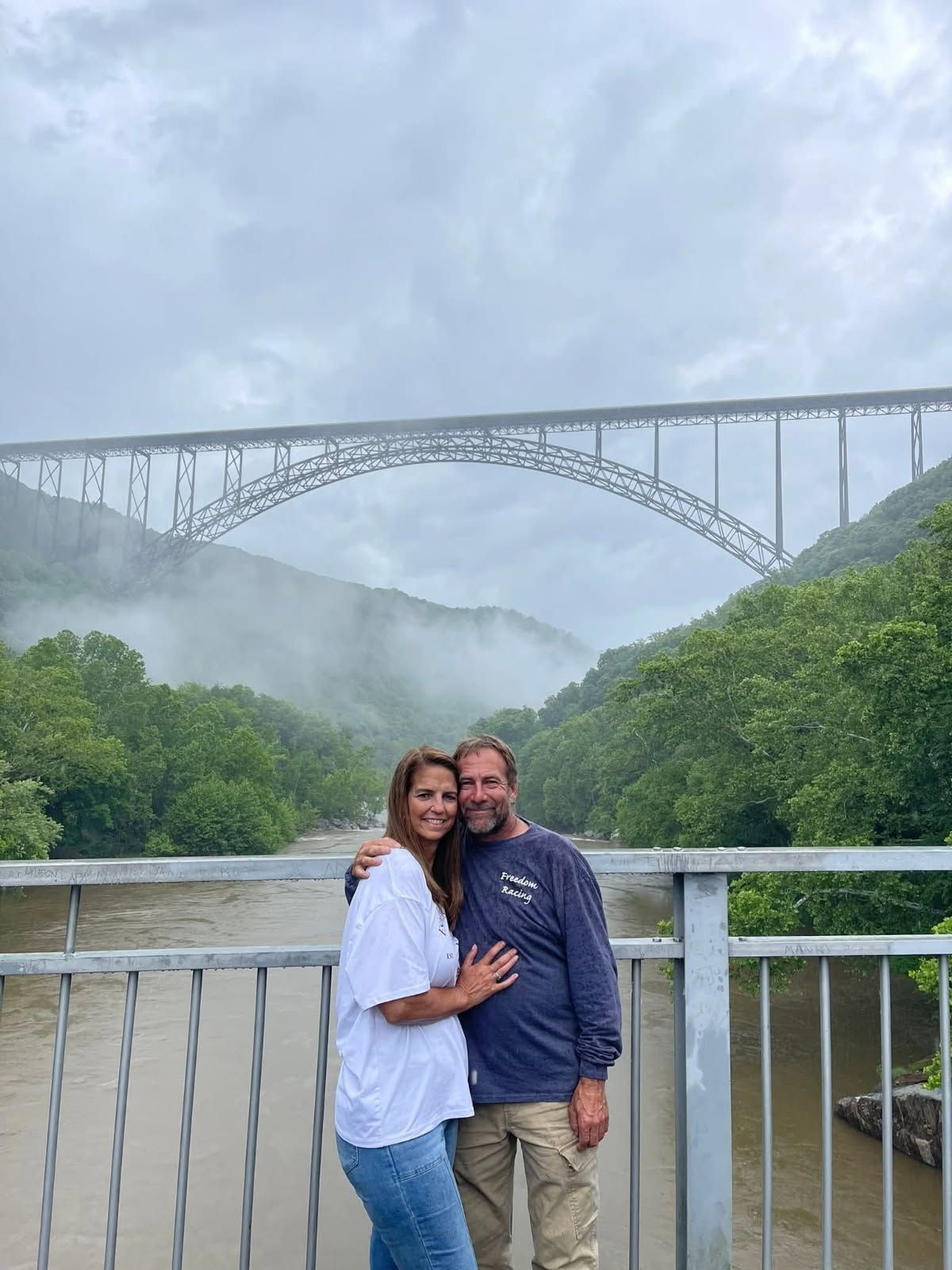 Couple posing on a bridge overlook with a large arched bridge and misty river valley behind them
