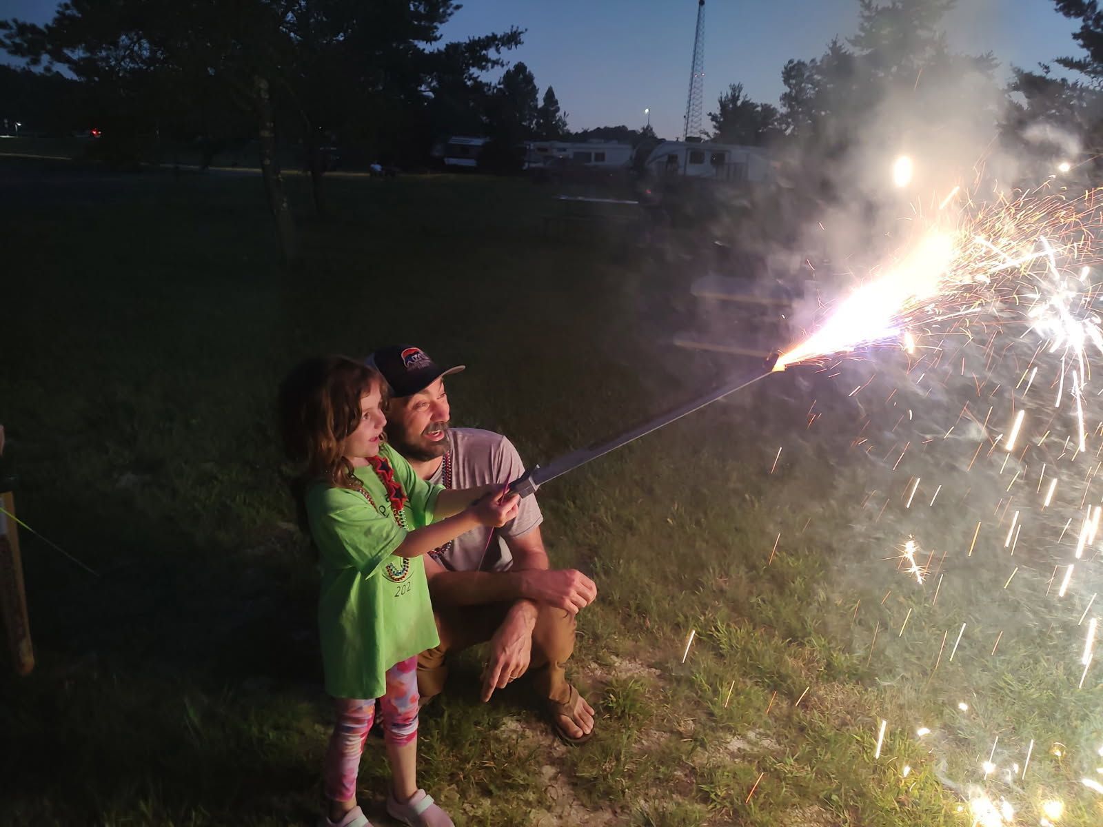 Two people sitting on grass watch a sparkling firework at dusk outdoors.