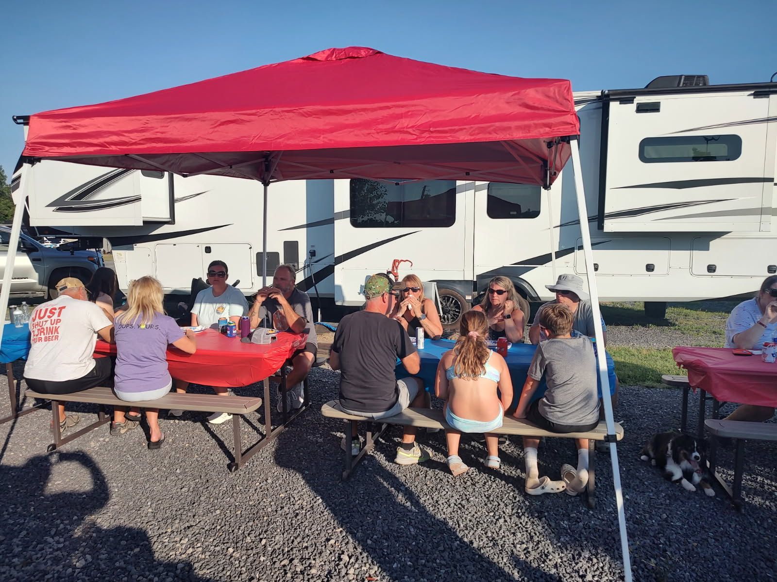 People gathered at picnic tables under a red canopy beside parked RVs on a gravel lot.