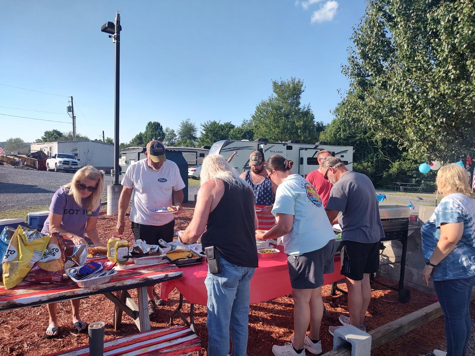People serving and eating food at outdoor picnic tables in a sunny park setting