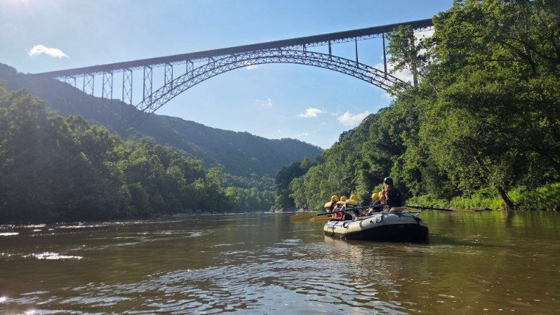 Rafting boat on a river beneath a large arched bridge, surrounded by green hills and blue sky