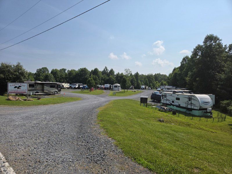 Gravel campground road with RVs and trailers parked along a grassy, tree-lined site under a blue sky