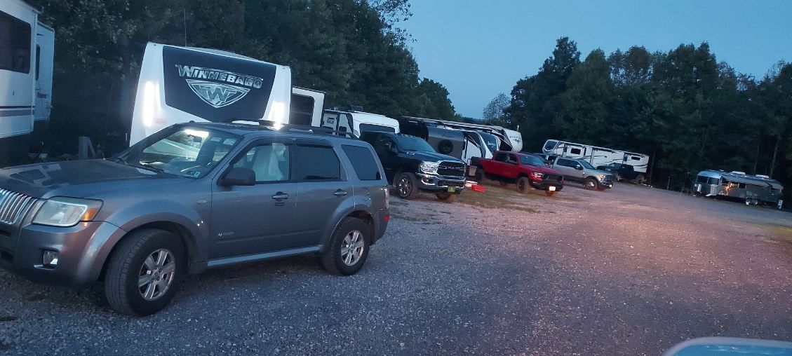 Cars parked at dusk in a gravel lot beside a lit roadside sign and trees.