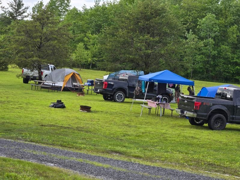Campsite with tents, SUVs, and a blue canopy on a grassy field beside trees