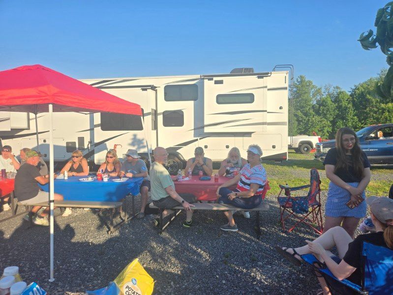 People sitting at picnic tables under a red canopy beside a large RV on a sunny day