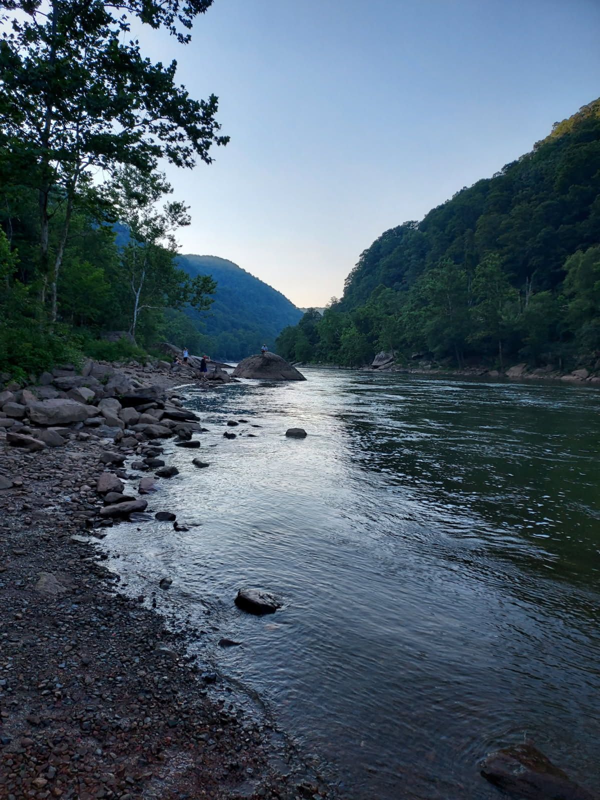 Rocky riverbank with calm water between forested hills at dusk