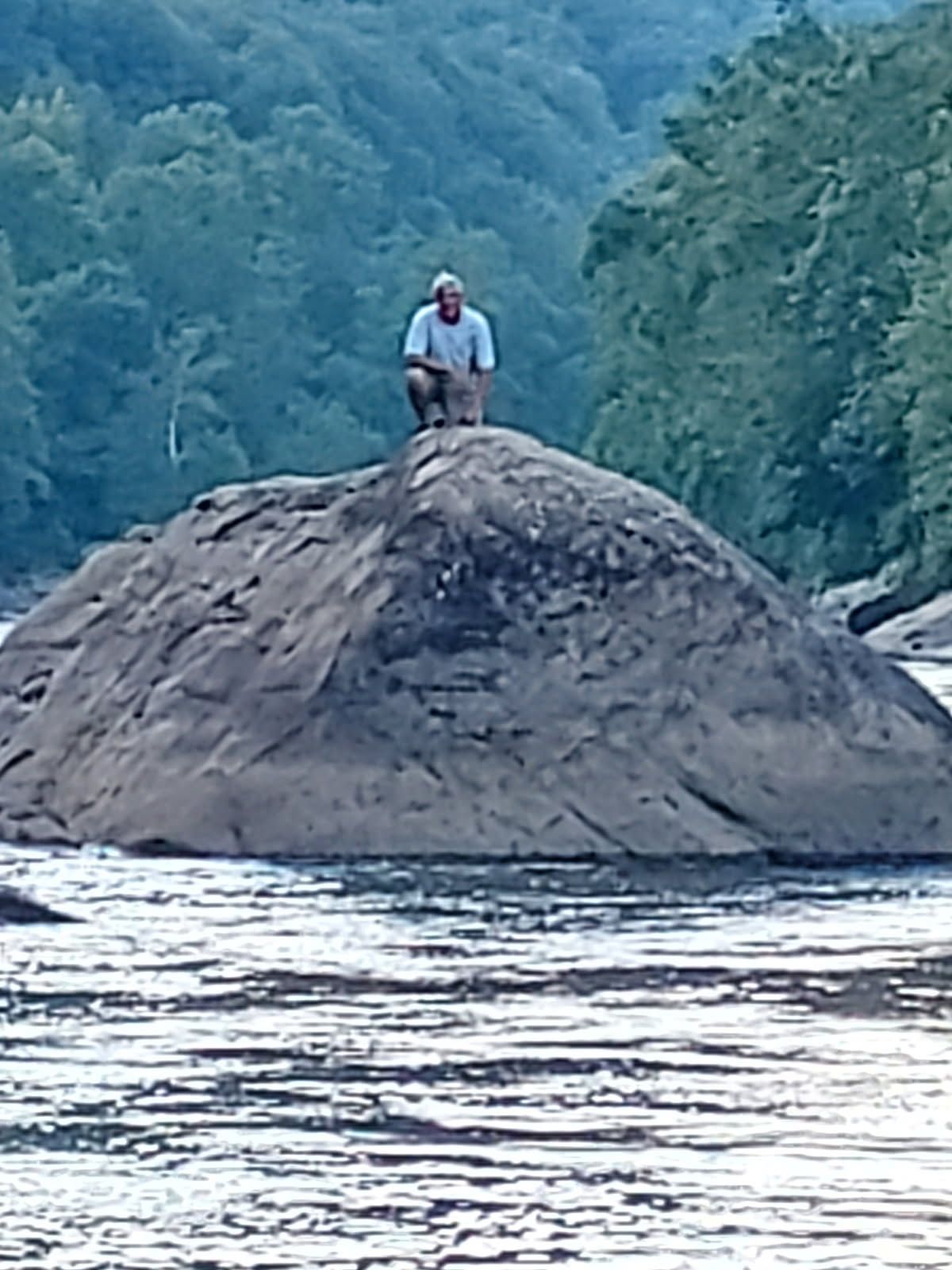 Person standing on a large rock in a river with forested hills in the background