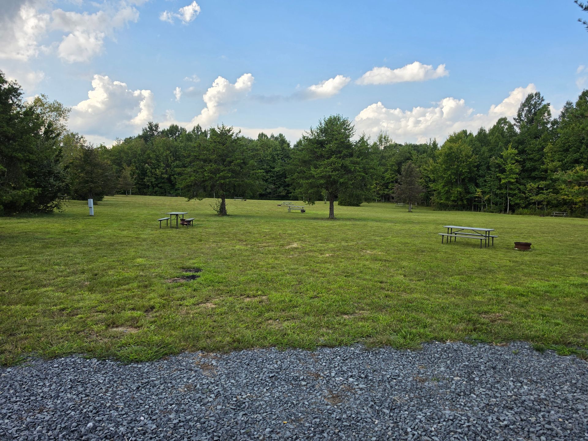 Grassy park with picnic tables, trees, and a gravel path under a blue sky with clouds