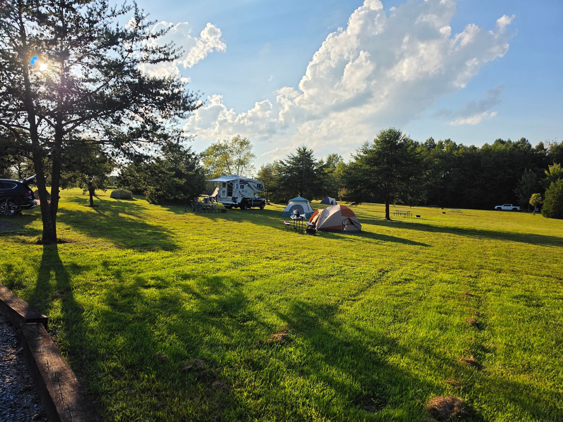 People relaxing on a sunlit grassy field with tents and a camper under a bright sky
