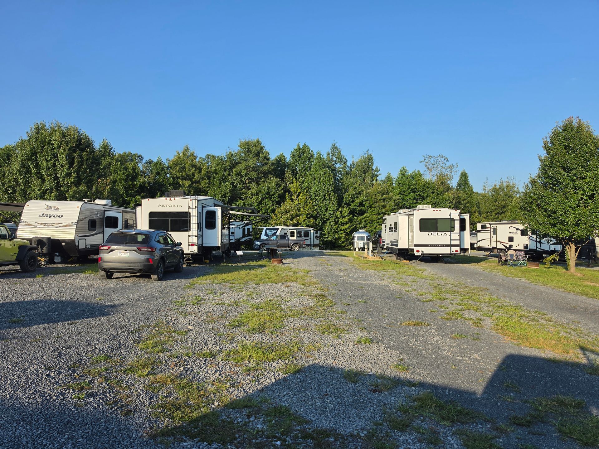 RV and camper parking area on gravel with cars, trees, and blue sky