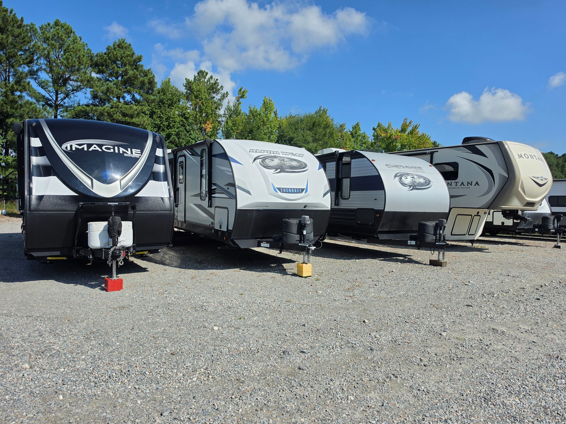 Row of white motorhomes parked on gravel beside trees under a blue sky