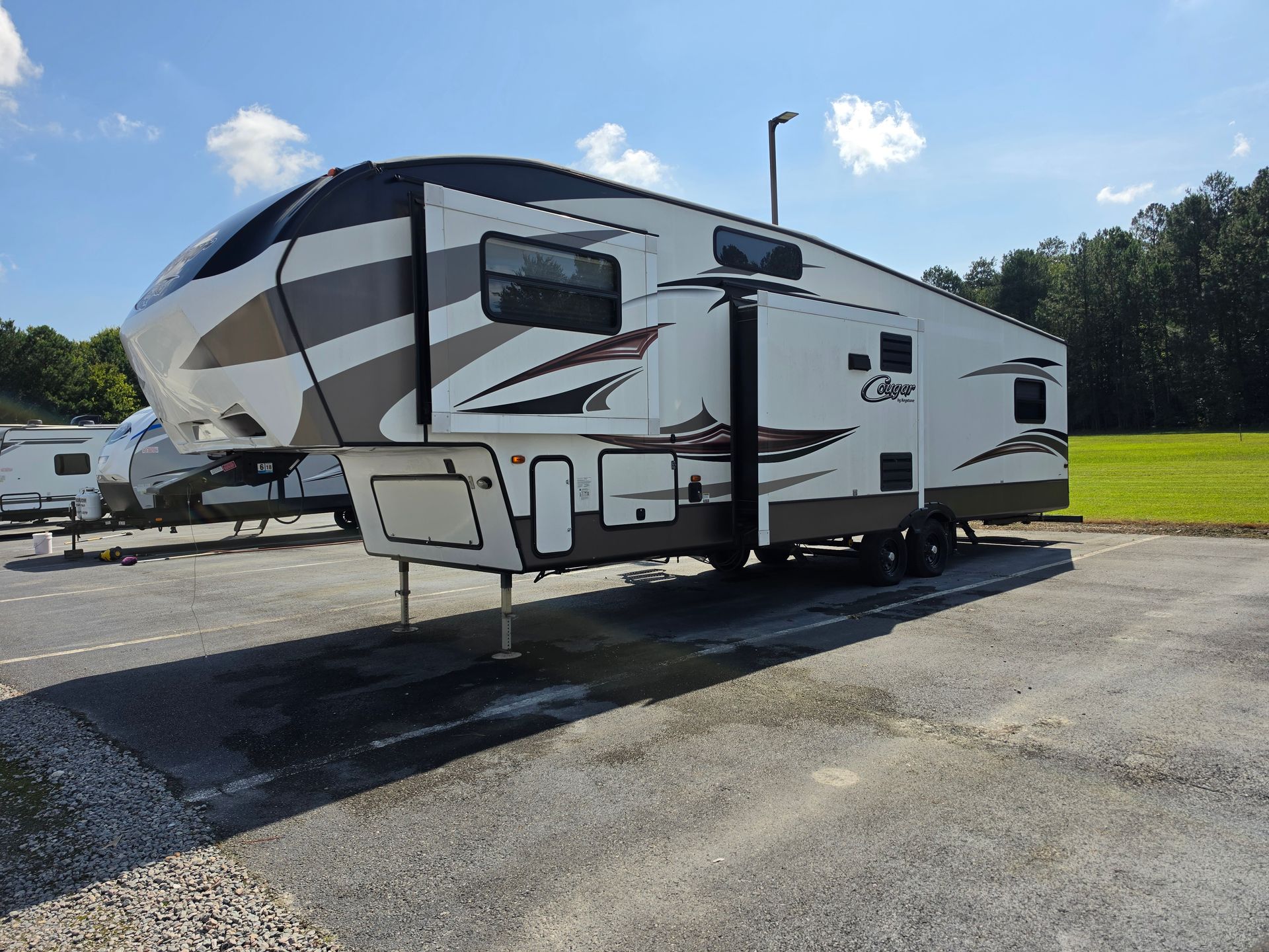 White and gray fifth-wheel RV parked on a paved lot under a blue sky, with trees and grass nearby
